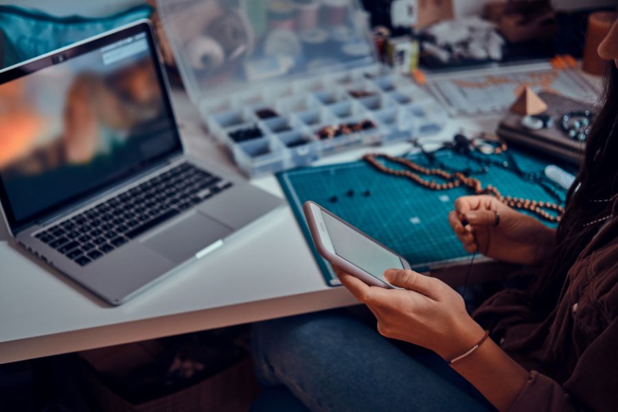 A person making beaded jewelry at a desk while looking at their smartphone. A person making beaded jewelry at a desk while looking at their smartphone.