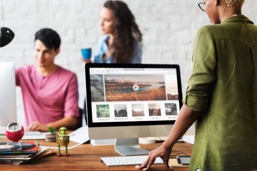 Three people working in a modern, creative office. A woman in the foreground is looking at a media website on a computer.