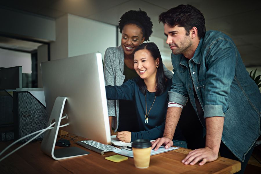 Three diverse colleagues smiling and working together on a computer in an office.