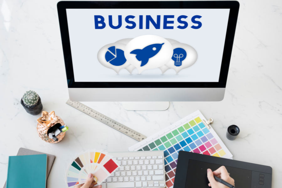 A graphic designer's desk with a computer screen that says "BUSINESS" and shows icons for a startup. A graphic designer's desk with a computer screen that says "BUSINESS" and shows icons for a startup.