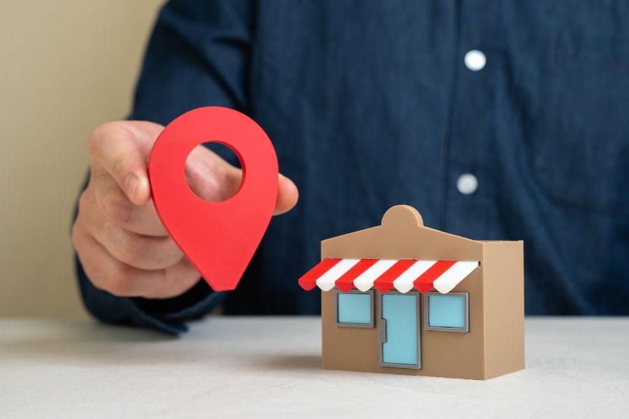 A person holding a large, red map pin icon next to a small model of a storefront. A person holding a large, red map pin icon next to a small model of a storefront.