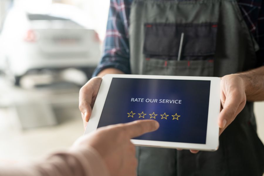 An auto mechanic holds a tablet that says "RATE OUR SERVICE," and a customer is about to tap one of the five stars to leave a rating. An auto mechanic holds a tablet that says "RATE OUR SERVICE," and a customer is about to tap one of the five stars to leave a rating.