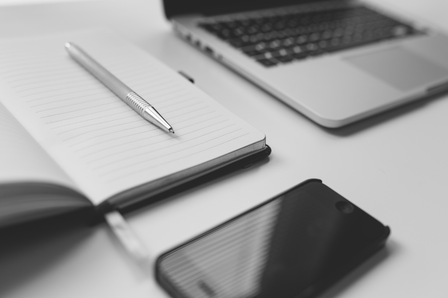 Black and white image of a notebook, a pen resting on it, and a smartphone beside them. Black and white image of a notebook, a pen resting on it, and a smartphone beside them.