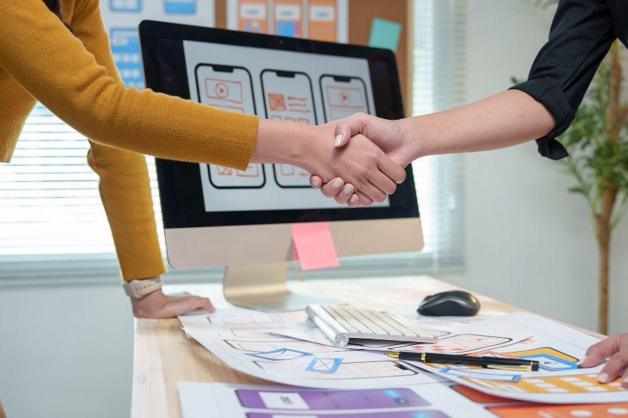 Two people shake hands over a desk in front of a computer showing mobile app designs.