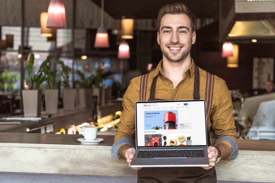 A smiling cafe owner in an apron holds up a laptop showing the website homepage. A smiling cafe owner in an apron holds up a laptop showing the website homepage.