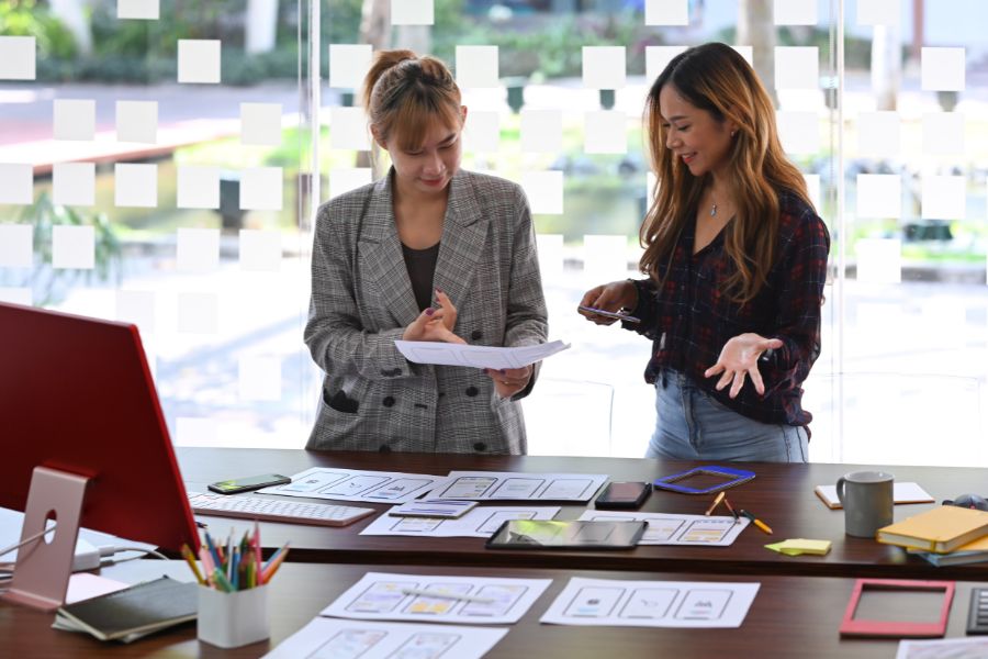 Two female designers collaborating over a desk covered with mobile app wireframes.