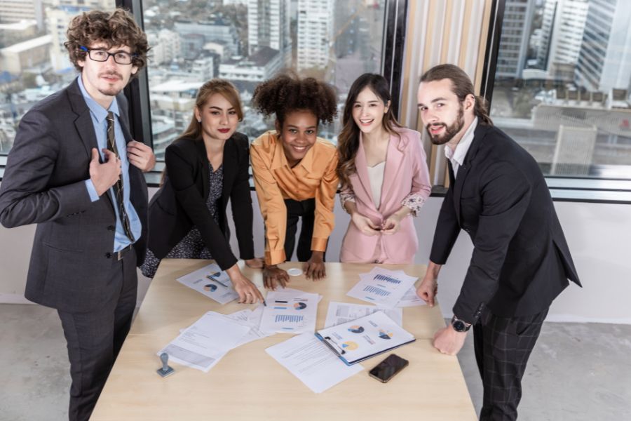 A diverse team of five business professionals poses for a photo around a table with charts in a high-rise office.